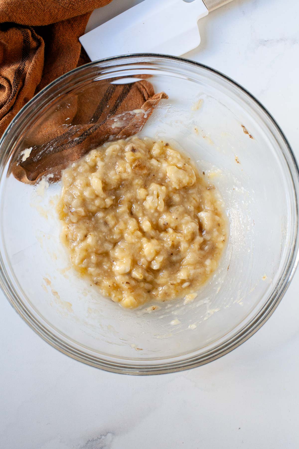 mashed banana in glass bowl with burnt orange dish cloth and white spatula