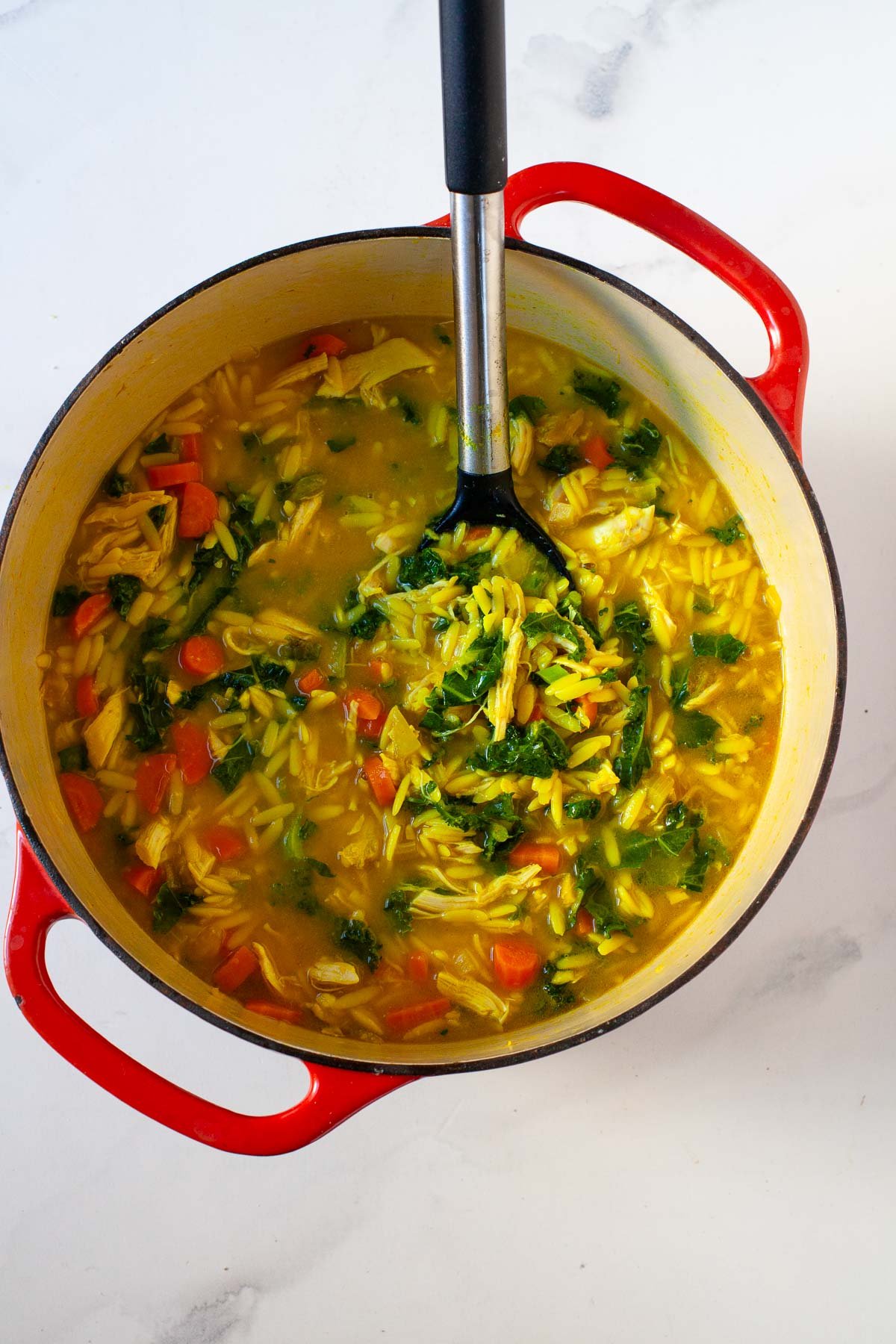 chicken orzo soup with kale, turmeric and ginger in red dutch oven with black mixing spoon