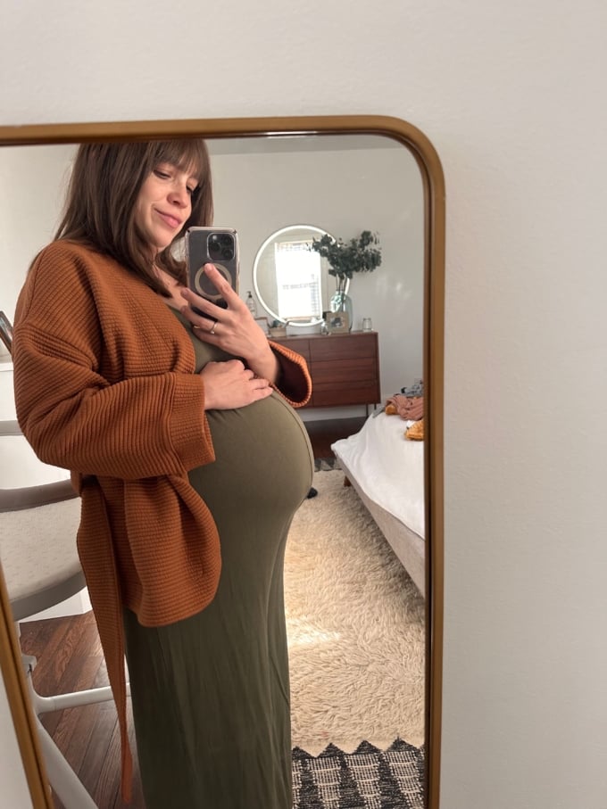 pregnant woman wearing green jumpsuit and sweater taking picture of herself in mirror